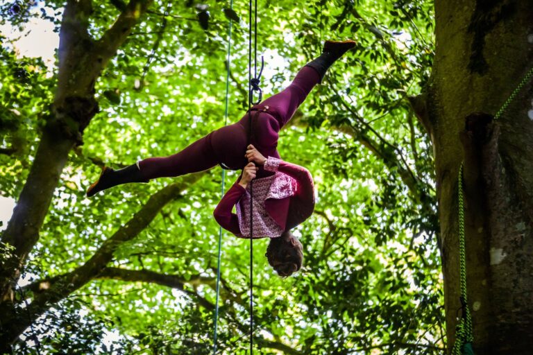 Une personne vêtue d'un vêtement marron exécute un saut aérien dans une impressionnante démonstration d'acrobatie, suspendue par des cordes entre des arbres verts dans un cadre arboré en plein air.