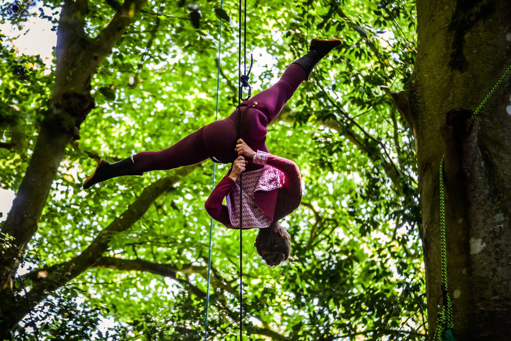 Une personne vêtue d'un vêtement marron exécute un saut aérien dans une impressionnante démonstration d'acrobatie, suspendue par des cordes entre des arbres verts dans un cadre arboré en plein air.