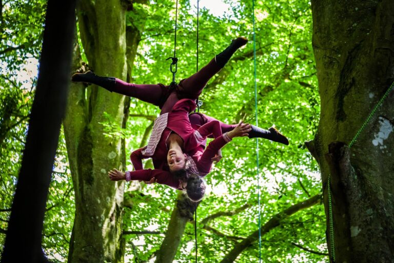 Deux acrobates vêtus de costumes bordeaux assortis exécutent un numéro d'acrobatie aérienne, suspendus la tête en bas parmi de grands arbres verts dans un cadre forestier paisible.