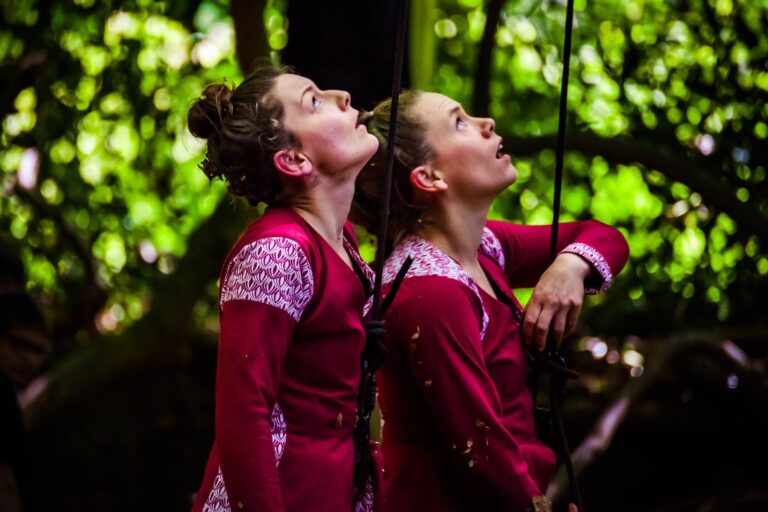 Deux femmes vêtues de tenues couleur framboise assorties se tiennent côte à côte, regardant vers le haut avec des expressions concentrées, incarnant la danse de la nature dans un cadre luxuriant et verdoyant en plein air.