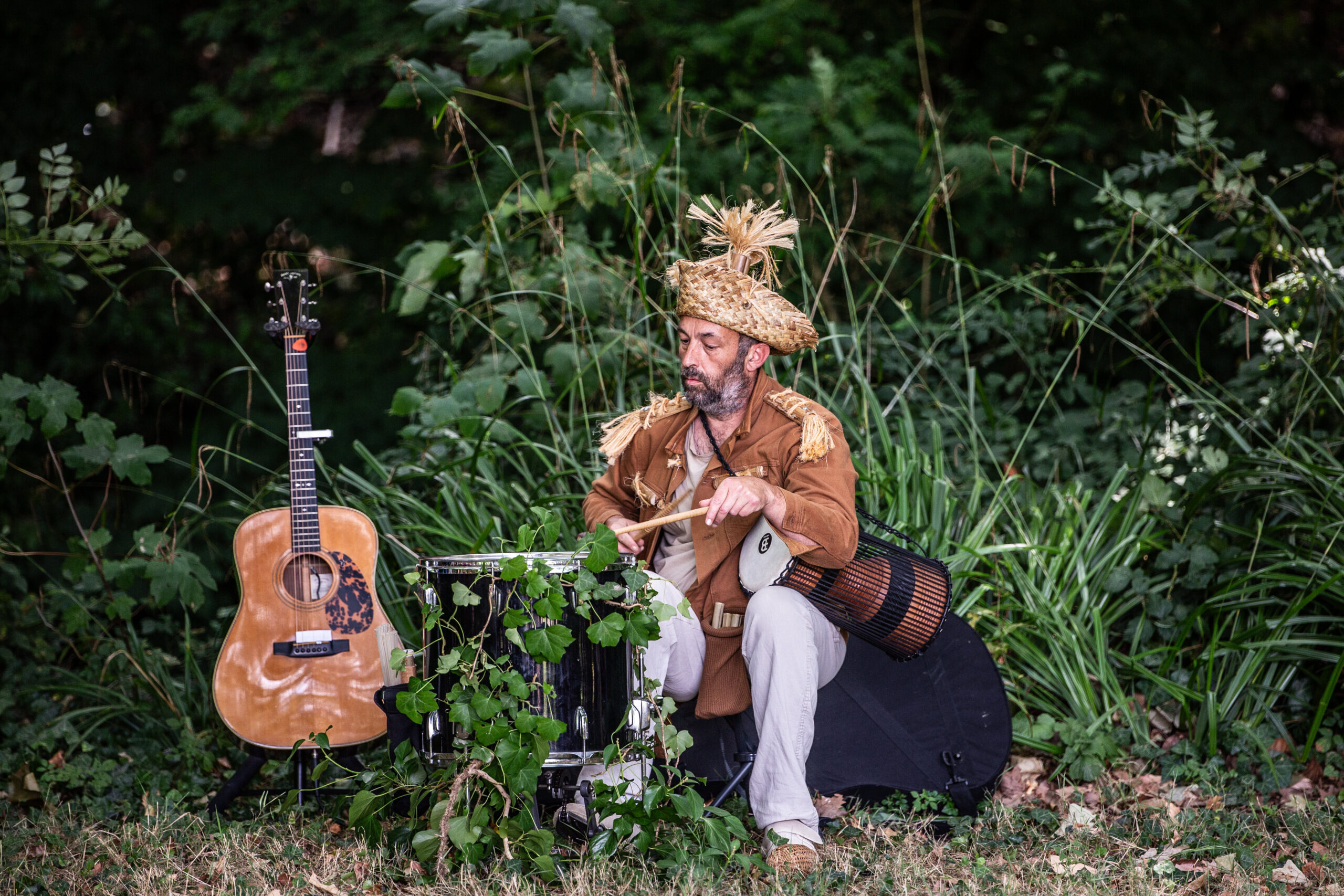 Un homme coiffé d'un chapeau décoratif orné de paille et vêtu d'un vêtement beige joue du tambour lors d'un spectacle en plein air, entouré de verdure, avec une guitare acoustique et d'autres tambours à proximité, sur l'herbe.