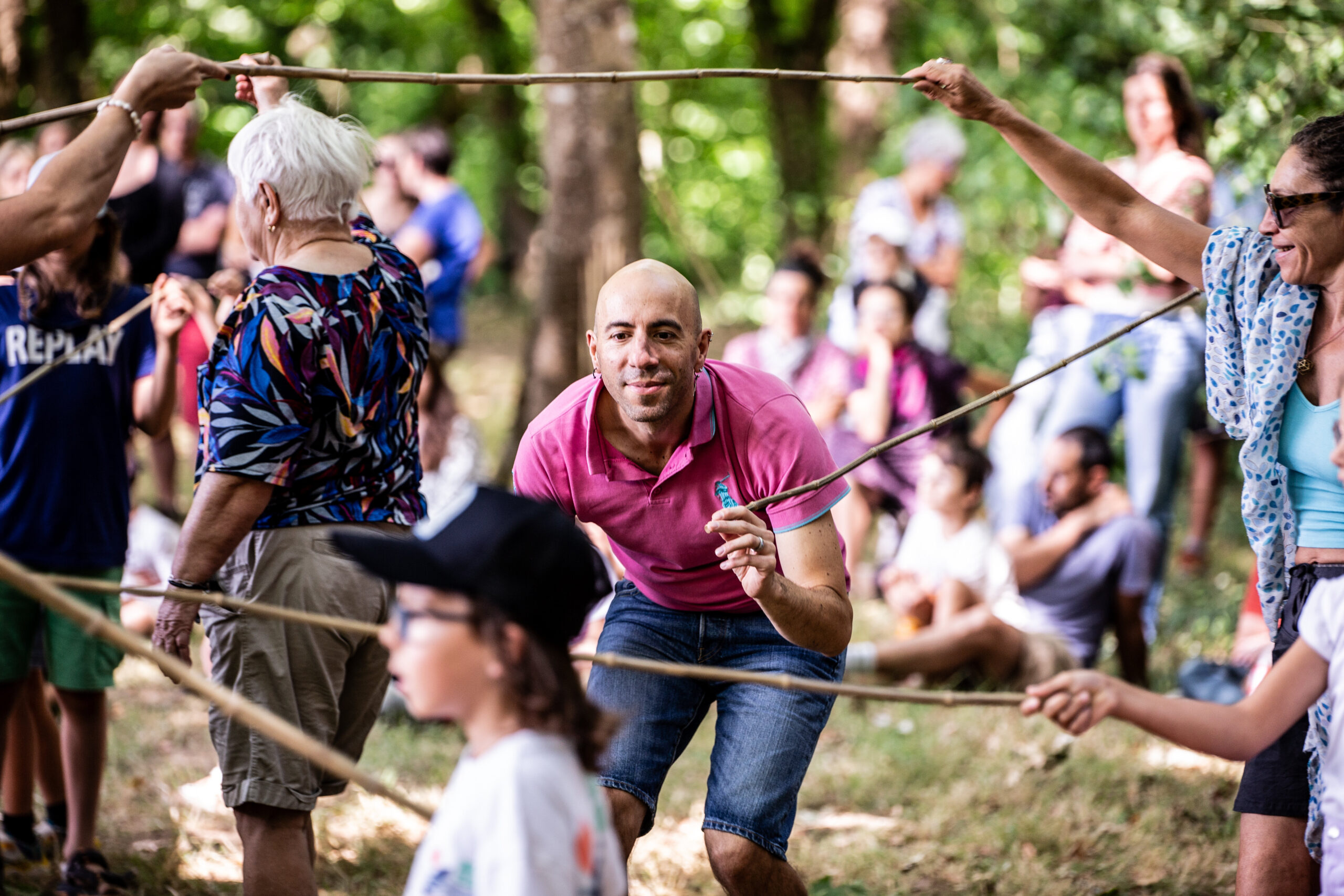 Un homme en chemise rose s'accroupit et sourit en se déplaçant sous des bâtons de bois tenus par d'autres lors d'un spectacle en plein air. Plusieurs personnes, jeunes et moins jeunes, participent ou observent ce spectacle en plein air dans un cadre boisé.