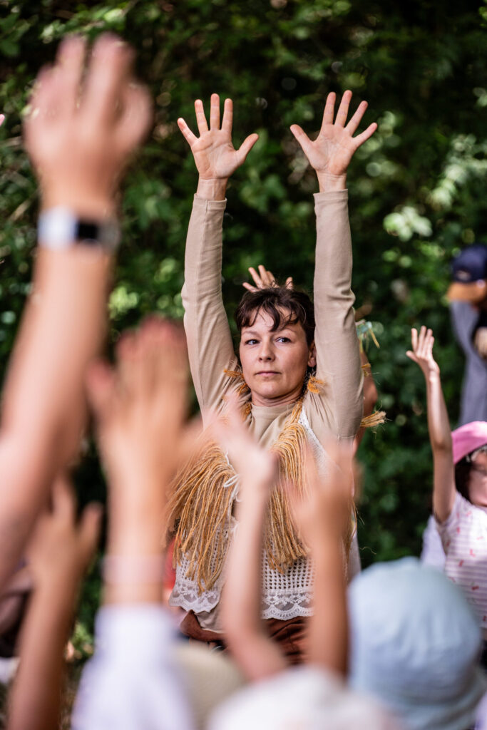 Une personne vêtue d'un haut beige à franges lève les bras au-dessus de sa tête lors d'un spectacle en plein air, entourée d'autres personnes qui font de même. L'arrière-plan est luxuriant et feuillu.