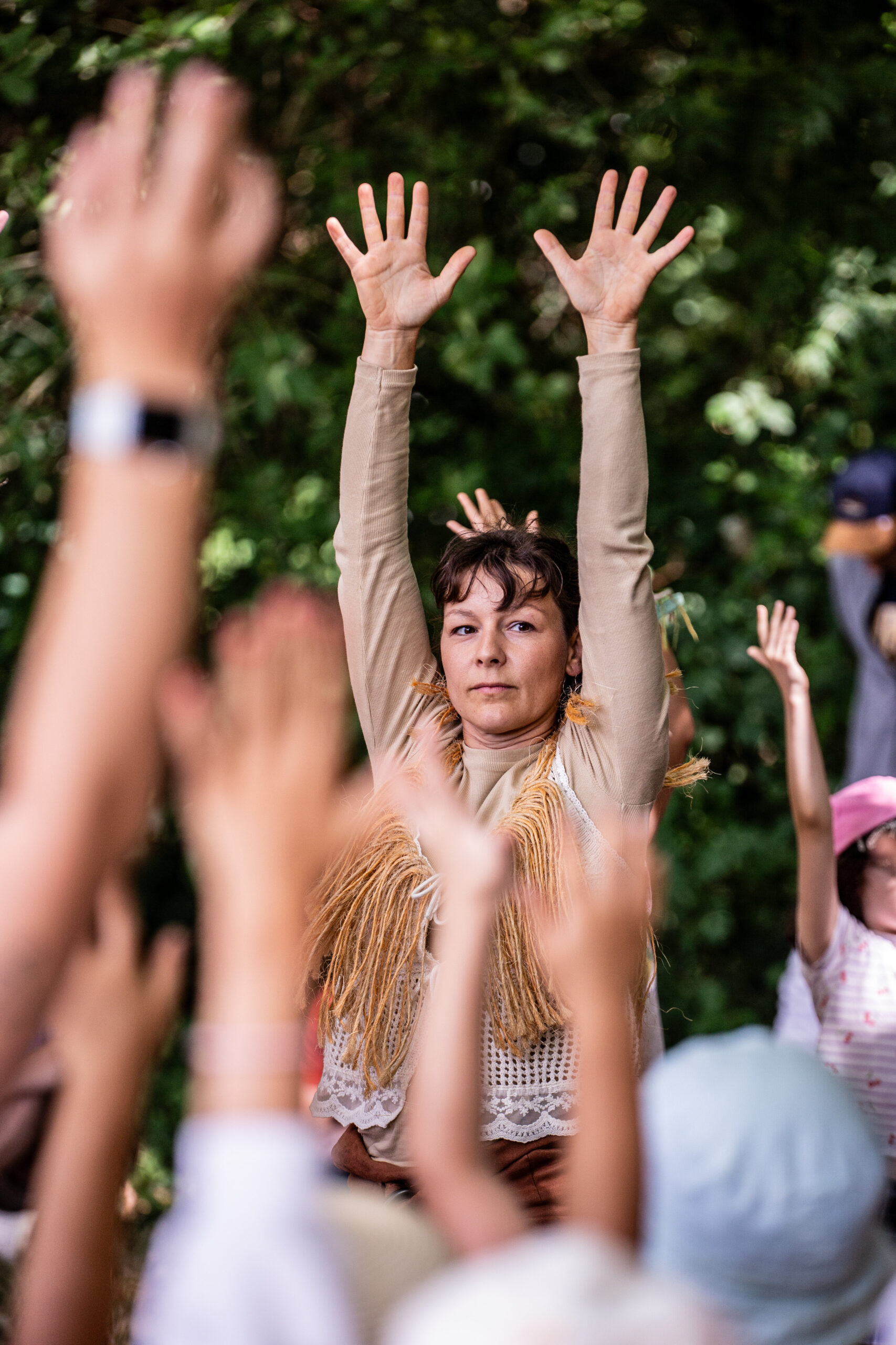 Une personne vêtue d'un haut beige à franges lève les bras au-dessus de sa tête lors d'un spectacle en plein air, entourée d'autres personnes qui font de même. L'arrière-plan est luxuriant et feuillu.