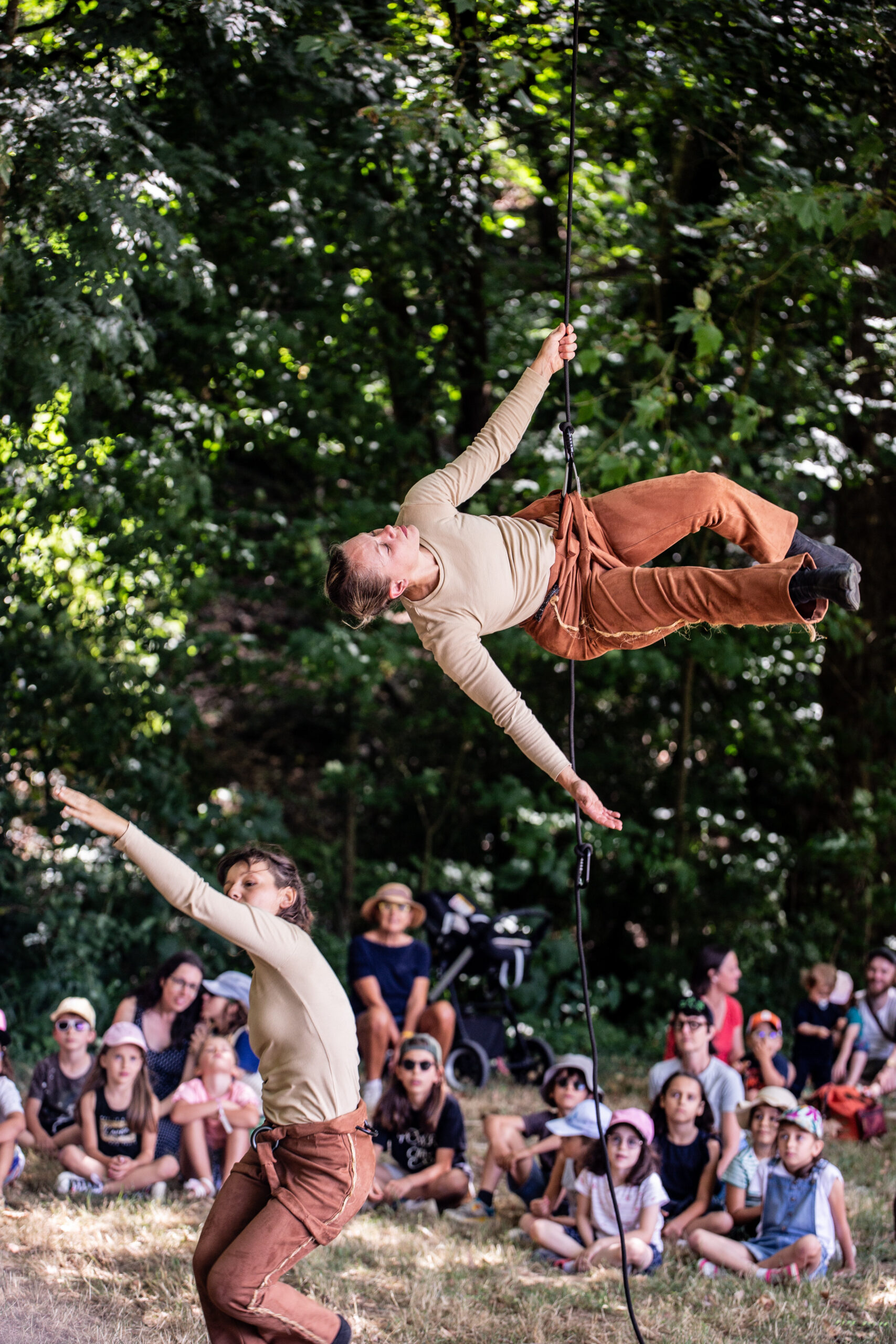 Deux artistes vêtus d'un haut beige et d'un pantalon marron présentent un spectacle en plein air, captivant un public assis avec leur numéro de corde acrobatique au milieu d'arbres verts, la lumière du soleil filtrant à travers les feuilles.