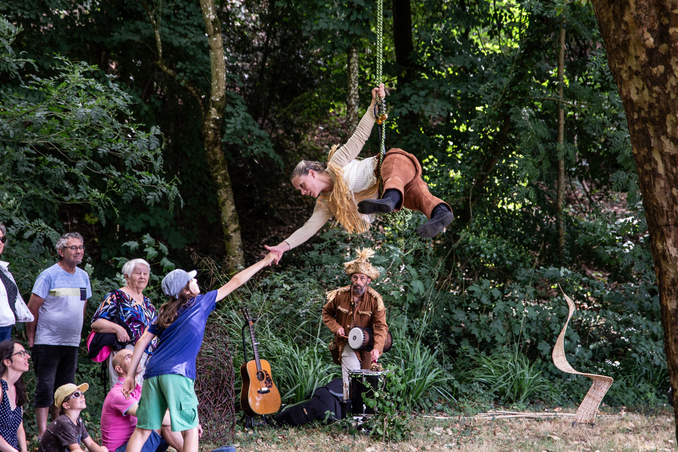 Un artiste sur une balançoire en corde tend la main à un jeune spectateur, sous le regard d'une petite foule dans un cadre boisé en plein air, lors d'un spectacle en plein air. Un musicien avec un tambour et une guitare est assis à proximité.