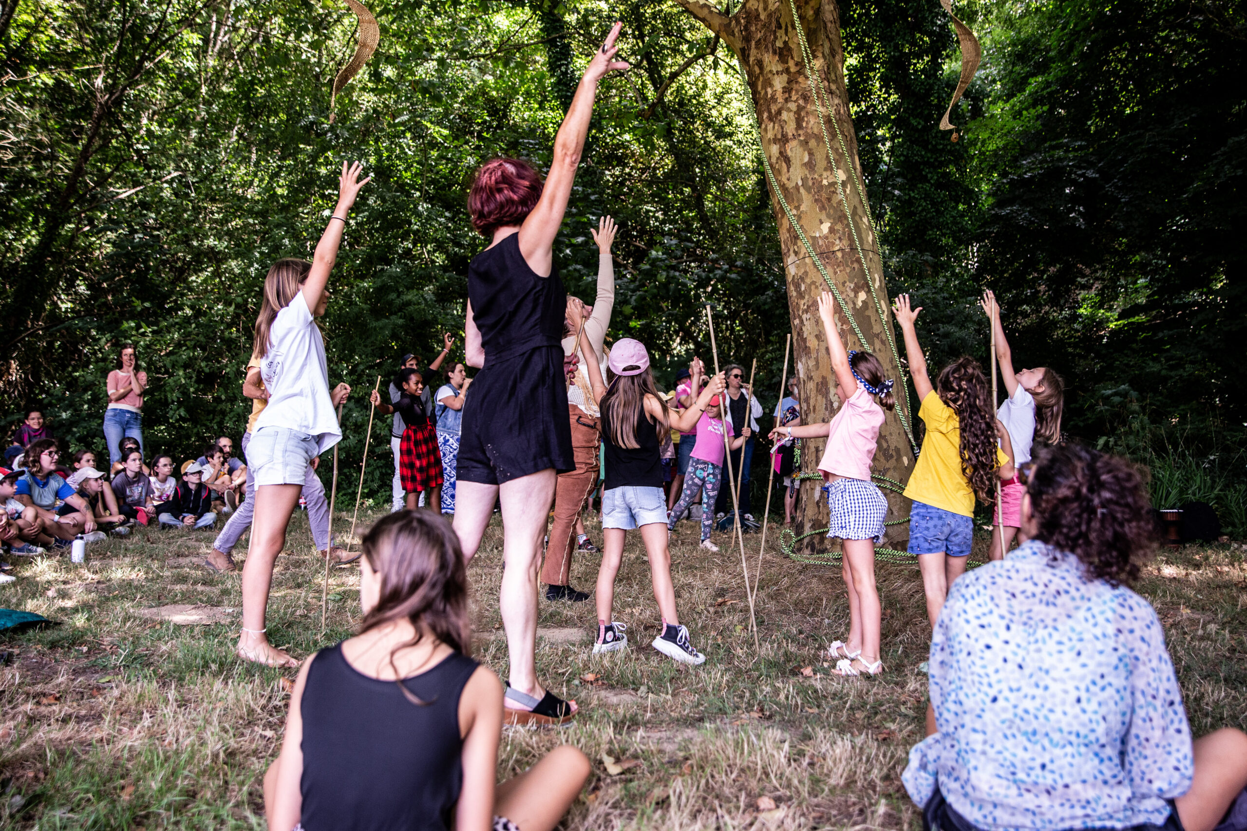 Un groupe d'enfants et d'adultes se tient dans un espace herbeux et boisé, les bras levés, et participe à un spectacle vivant en plein air, tandis que d'autres s'assoient et observent en arrière-plan.