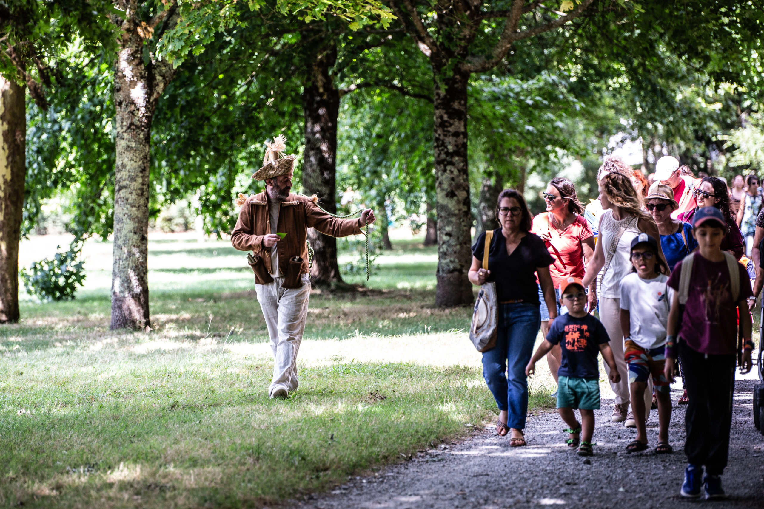 Une personne vêtue d'un costume en matériaux naturels s'approche d'un groupe de personnes, adultes et enfants, lors d'un spectacle en plein air le long d'un chemin bordé d'arbres dans un parc ensoleillé.