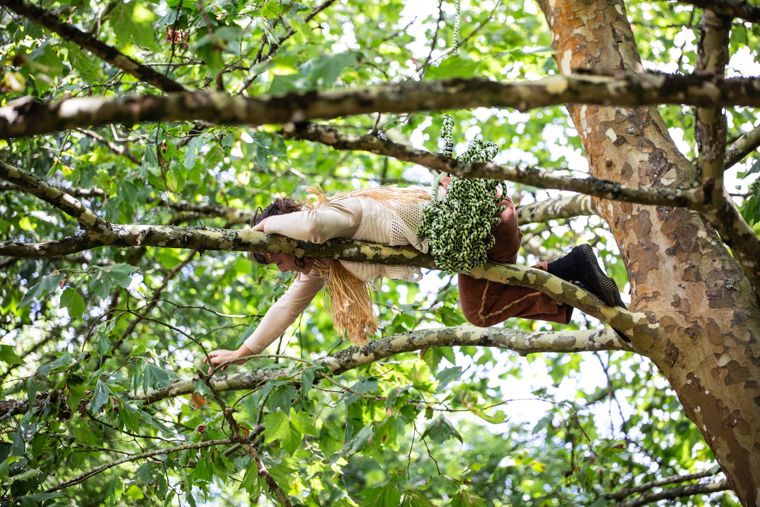Une personne est allongée sur une branche d'arbre lors d'un spectacle en plein air, tendant un bras vers l'avant. Elle porte un pull beige, un pantalon marron et des bottes noires. Des fleurs blanches reposent à côté d'elle parmi des feuilles vertes.