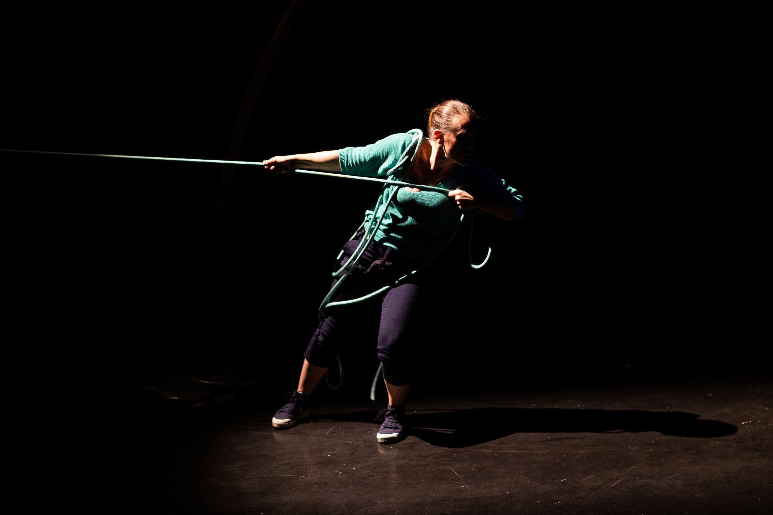 Une personne est éclairée sur une scène sombre et exécute une danse suspendue en tirant sur une longue corde qui s'enroule autour de son corps.