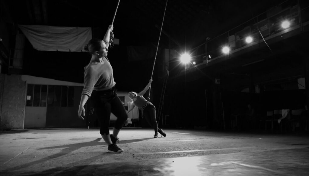 Deux personnes se tiennent à des cordes et se penchent en arrière pour exécuter une danse encordée expressive sur une scène faiblement éclairée, les lumières du plafond illuminant l'espace vide du public.
