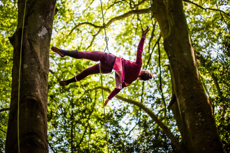 Une personne en tenue rouge fait de l'acrobatie arbre, suspendue à un harnais entre deux grands arbres dans une forêt, encadrée par un feuillage vert luxuriant et la lumière du soleil.
