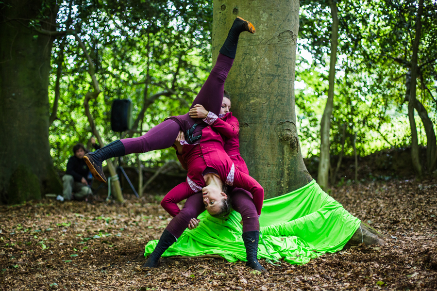 Deux artistes en tenue bordeaux assortie font une démonstration d'acrobatie cirque près d'un arbre dans une zone boisée, avec un tissu vert vif drapé à sa base.