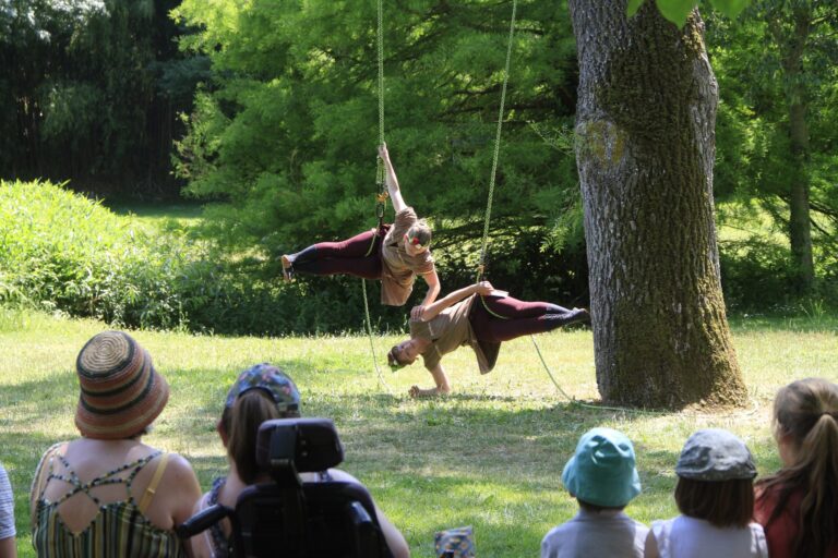 Deux artistes aériens créent un spectacle époustouflant, de danse encordée suspendues horizontalement à des cordes attachées à un arbre, sous le regard d'un public en plein air, dans l'herbe, par une journée ensoleillée.