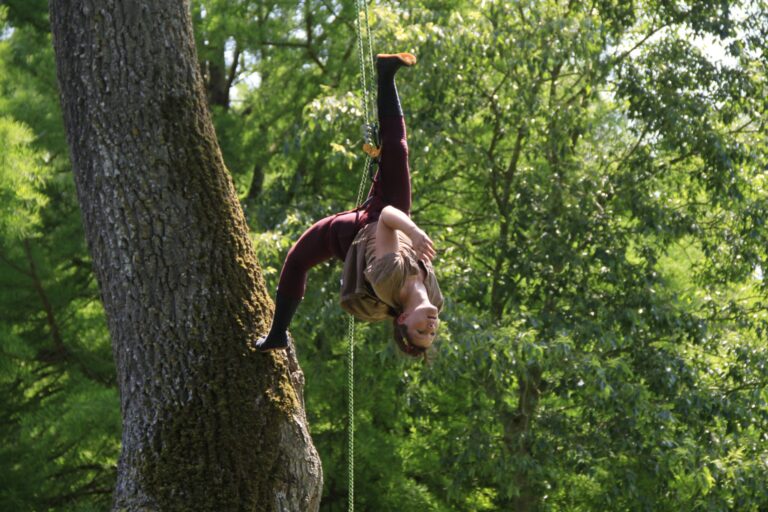 Une personne exécute une pose acrobatique aérienne, suspendue à l'envers par une jambe à une corde attachée à un grand arbre, incarnant la grâce de la danse acrobatique dans la nature, dans un cadre forestier paisible.