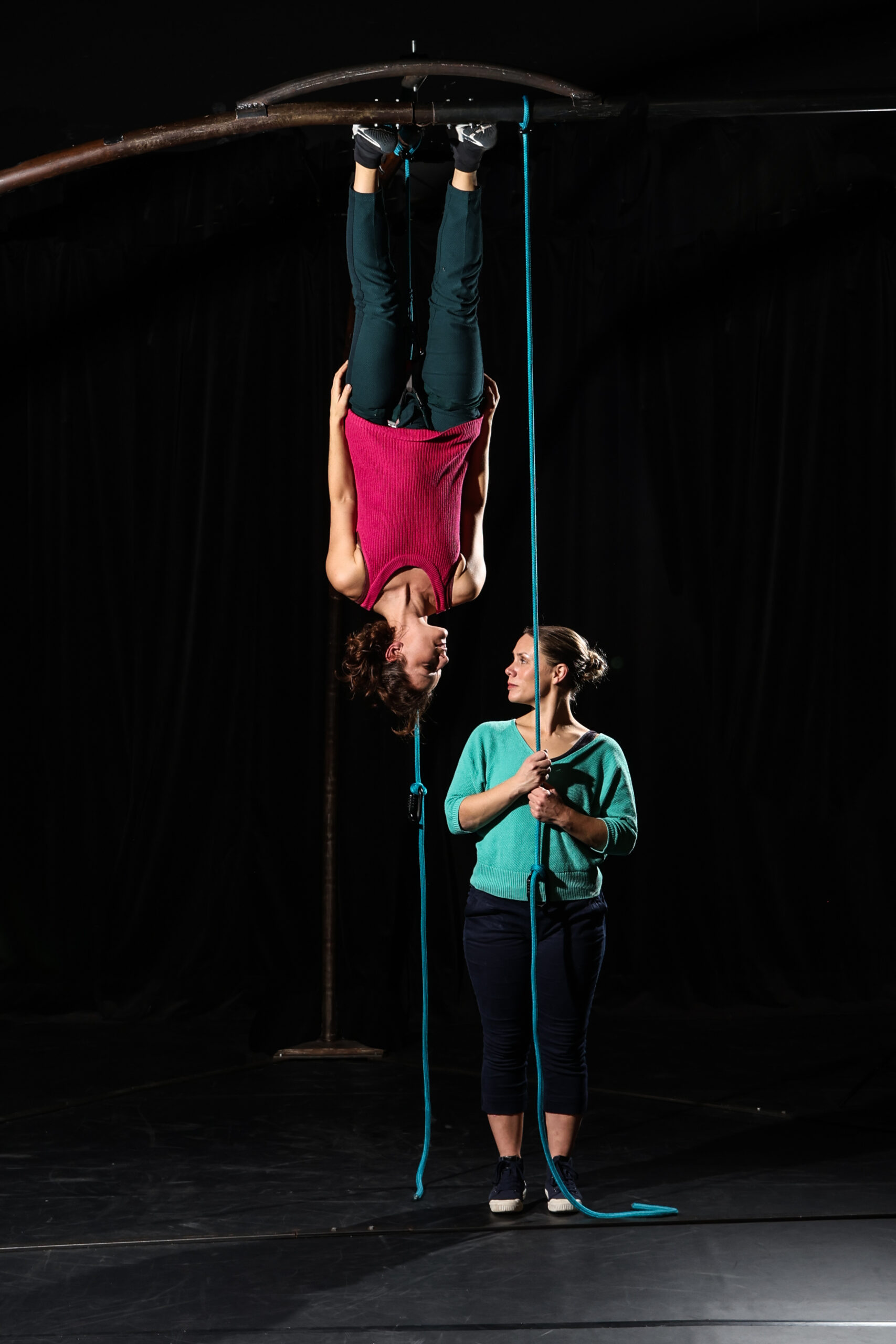 Une personne exécute une danse de cirque, suspendue par les jambes à un appareil aérien, tandis qu'une autre se tient à proximité en tenant une corde bleue, toutes deux sur une scène faiblement éclairée.