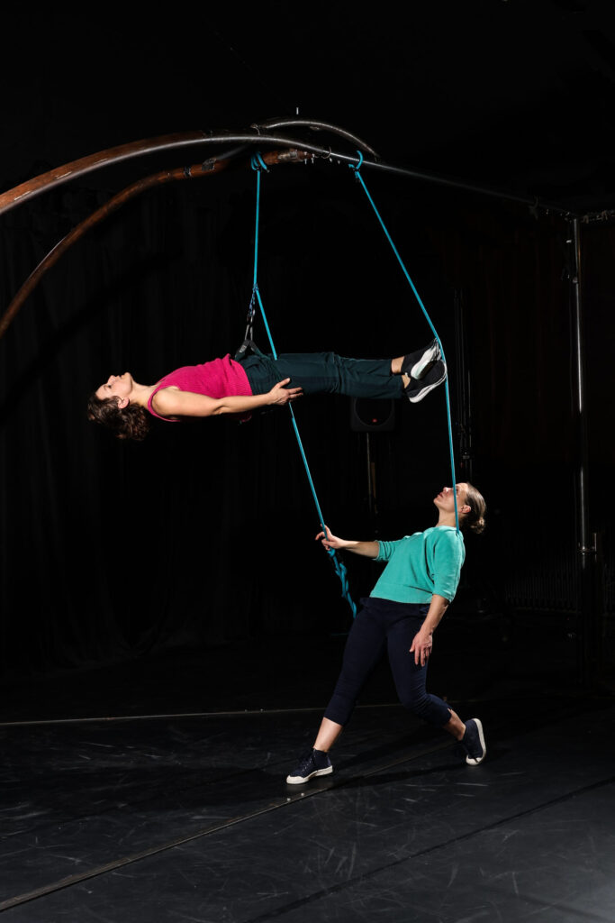 Deux personnes exécutent un numéro de danse acrobatique ; l'une est suspendue horizontalement par des cordes, tandis que l'autre les soutient par le bas sur une scène sombre.
