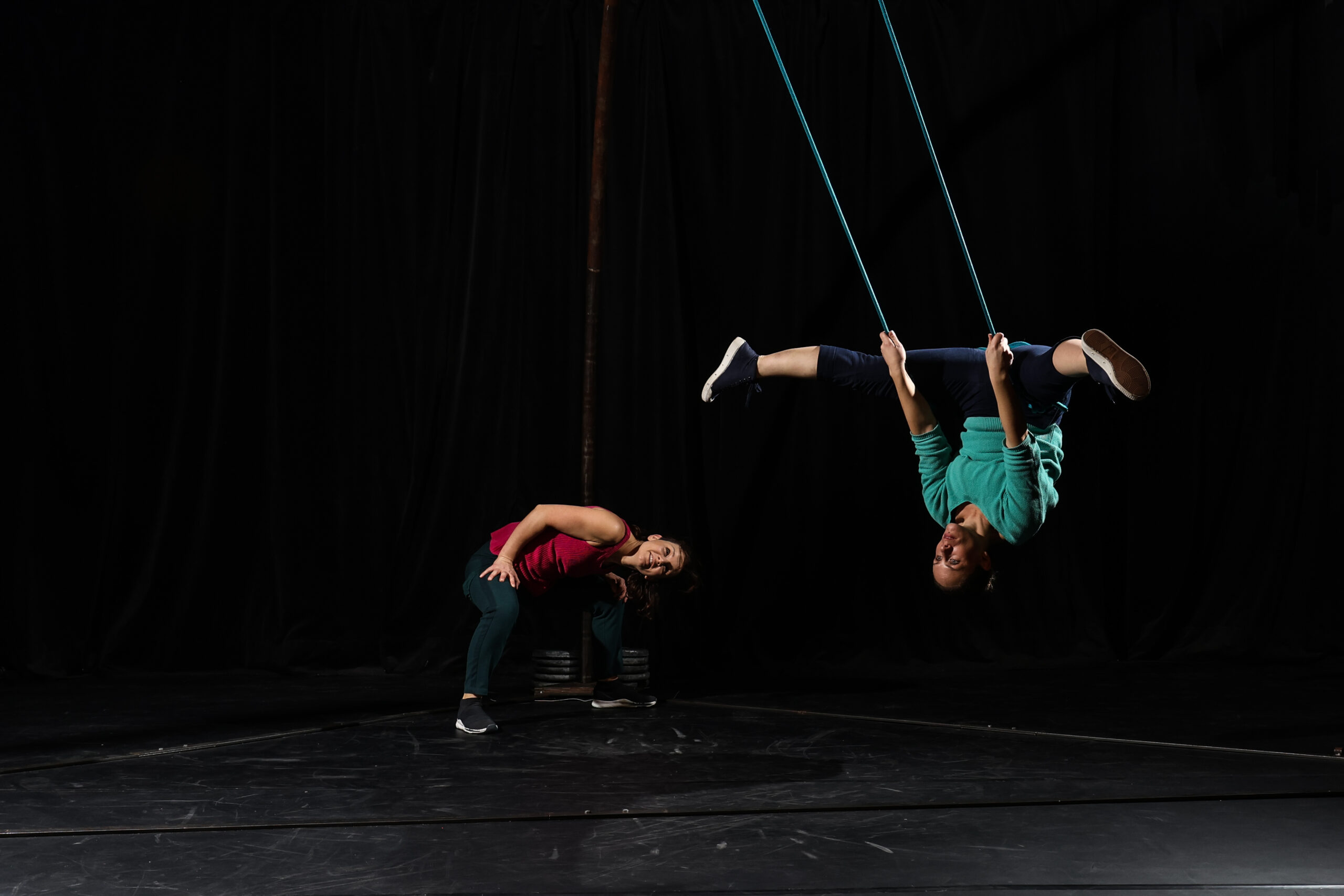 Deux personnes font de l'acrobatie sur scène ; l'une est suspendue à des cordes, la tête en bas, tandis que l'autre se tient à proximité, penchée vers l'avant. Des rideaux noirs servent de toile de fond.