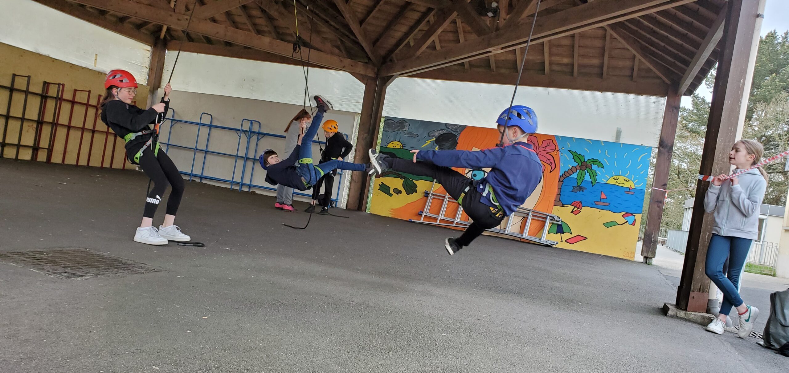 Quatre enfants portant des casques et des harnais se balancent sur des cordes sous un abri en bois dans un établissement scolaire, tandis qu'un autre enfant observe à proximité. Une peinture murale colorée est visible à l'arrière-plan.