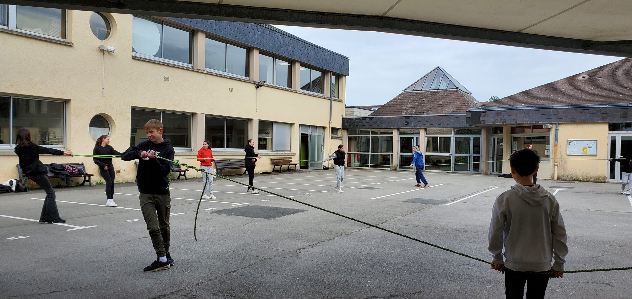 Un groupe d'élèves dans la cour d'une école, suivent un atelier danse au sol, deux tenant les extrémités d'une corde au sein d'un groupe.