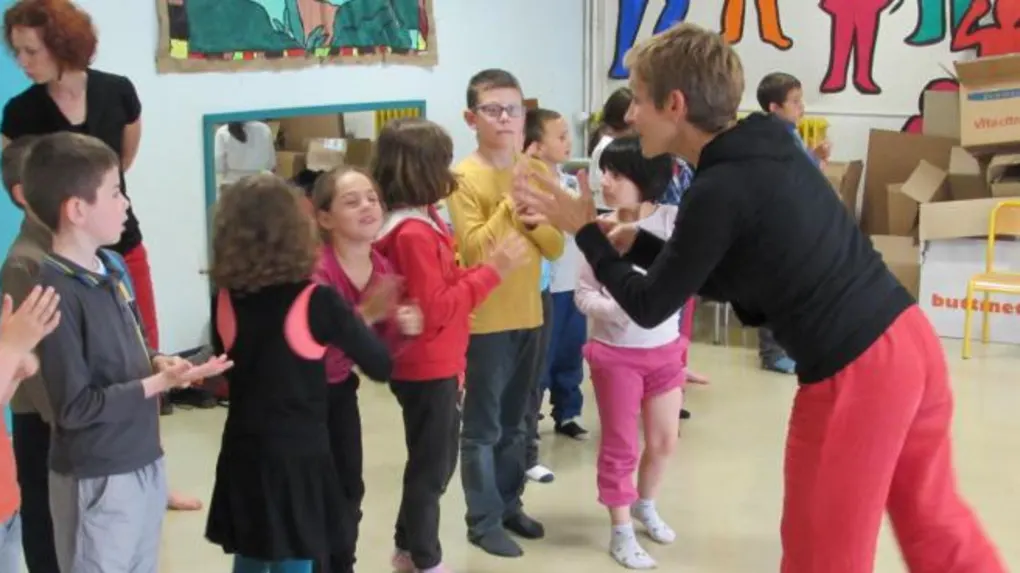 Un groupe d'enfants se tient dans une salle de classe de l'école Jules-Ferry, participant à une activité de danse interactive dirigée par un adulte en pantalon rouge. Des œuvres d'art colorées décorent les murs.