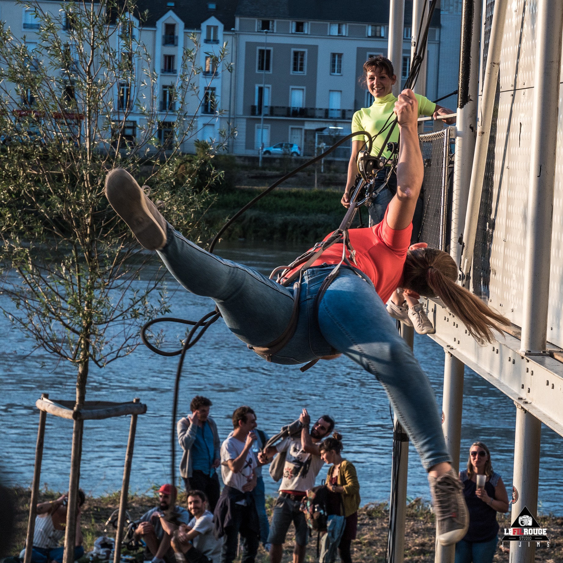 Une femme dans un harnais se balance d'une structure au-dessus de la Loire, tandis qu'une personne souriante la surveille et qu'un groupe de spectateurs la regarde d'en bas.