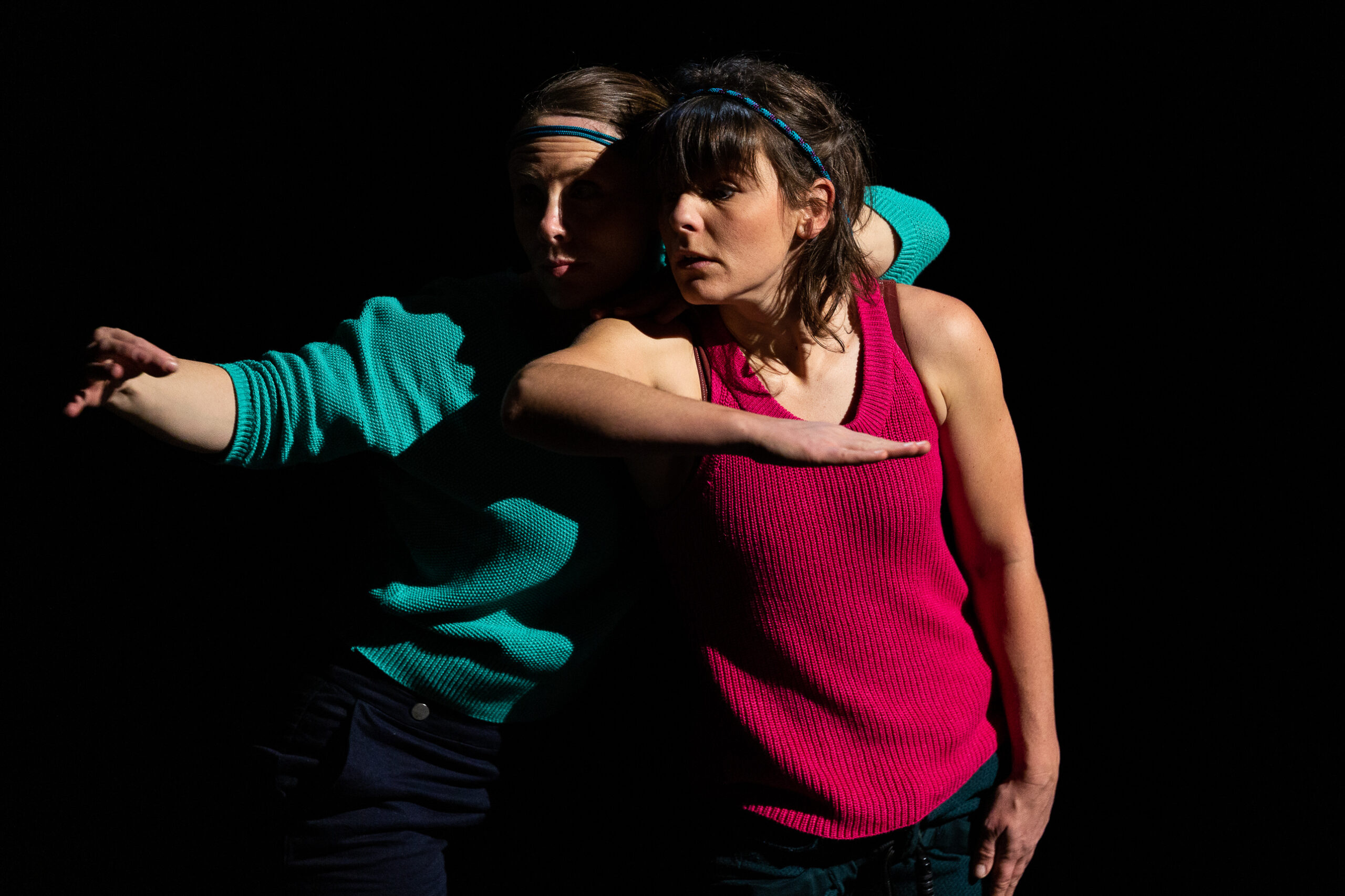 Deux femmes posent l'une près de l'autre dans un style de danse contemporaine saisissant, l'une vêtue d'un pull sarcelle et l'autre d'un débardeur rose, toutes deux étendant leurs bras de manière spectaculaire sur un fond noir.
