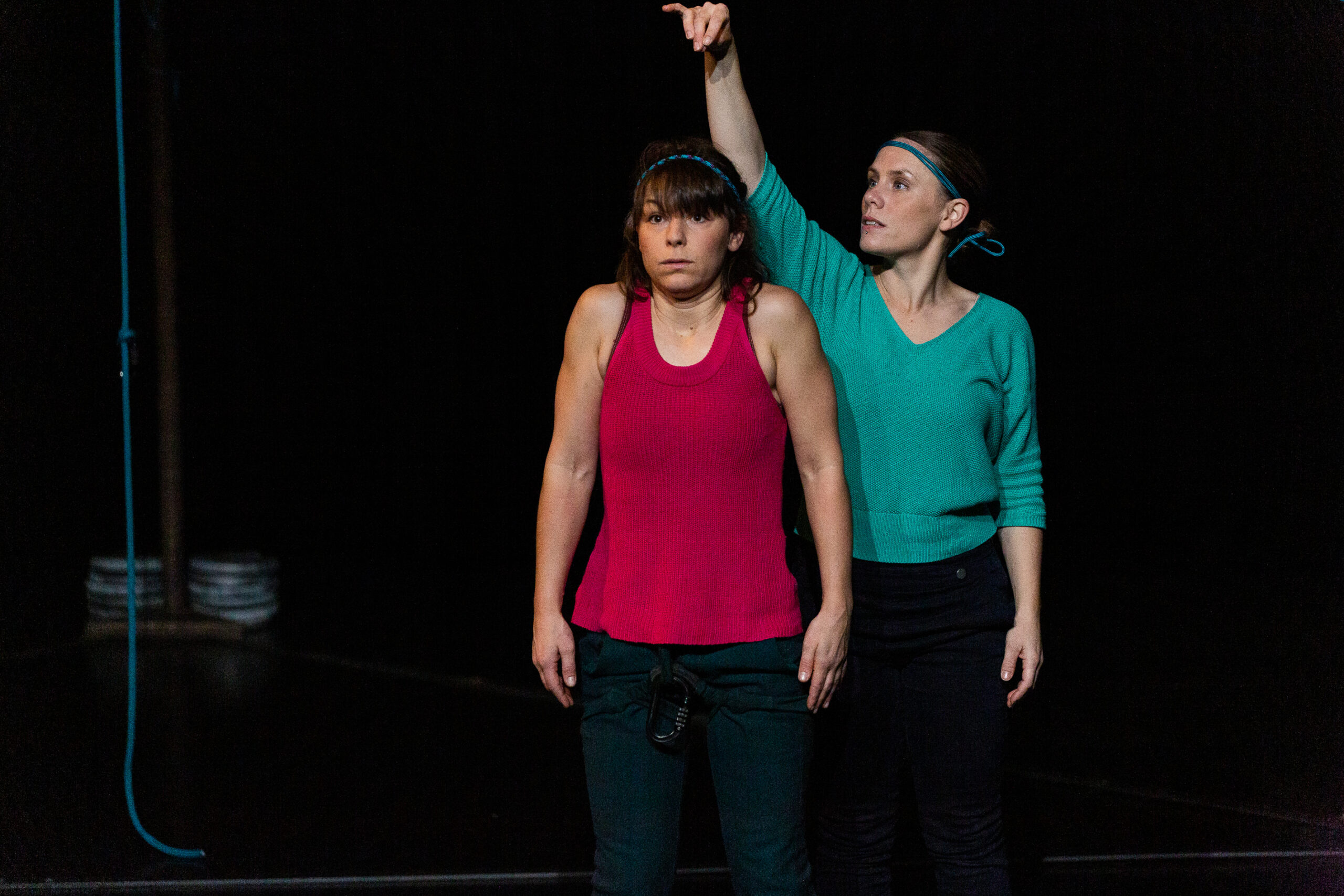 Deux femmes se tiennent sur une scène sombre dans un moment intense de danse contemporaine. La femme à l'avant porte un débardeur rose et semble mal à l'aise, tandis que la femme derrière elle, en vert, pointe vers le haut avec une expression sérieuse.