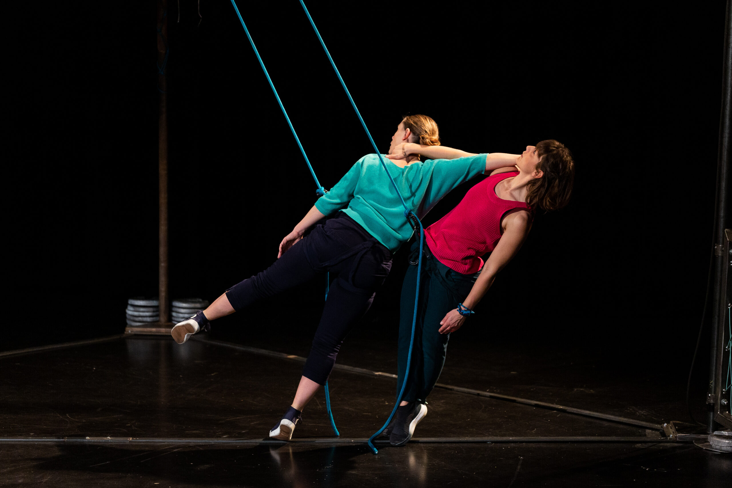 Deux femmes exécutent une danse escalade hypnotique, appuyées l'une contre l'autre et suspendues par des cordes bleues sur une scène sombre.