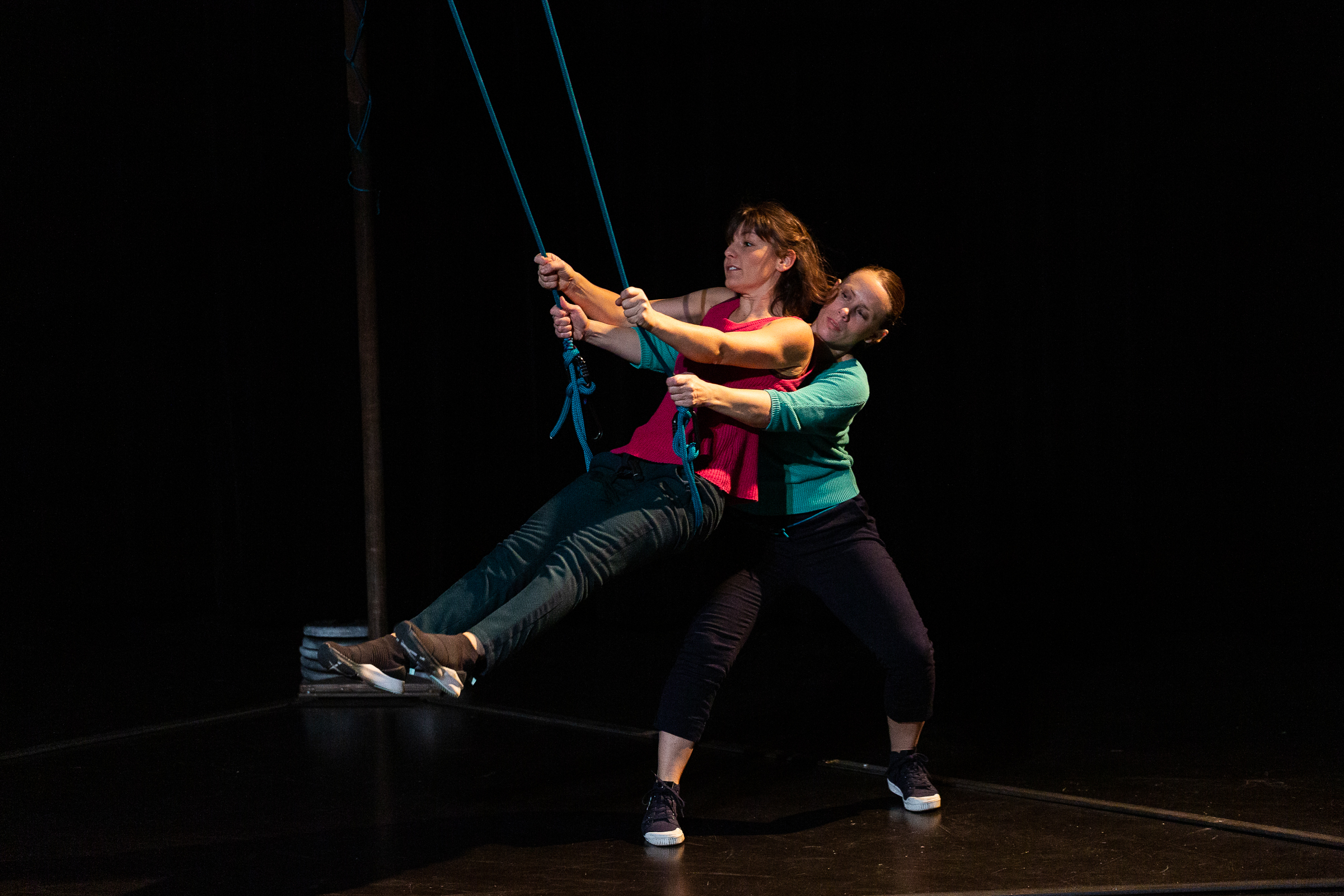 Deux femmes exécutent une danse verticale avec des cordes bleues sur scène. L'une se balance tandis que l'autre la soutient par derrière. Toutes deux portent des vêtements décontractés sur un fond sombre.