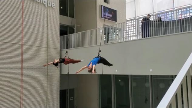 Deux personnes harnachées exécutent un spectacle de danse aérienne à couper le souffle, suspendues horizontalement dans les airs à l'intérieur d'un bâtiment moderne dont les balustrades et les balcons sont visibles à l'arrière-plan.