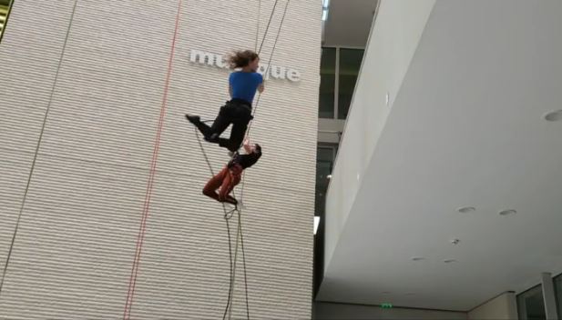 Deux personnes portant des harnais effectuent un spectacle aérien sur des cordes à l'intérieur d'un bâtiment moderne avec de hauts plafonds et un mur rayé.
