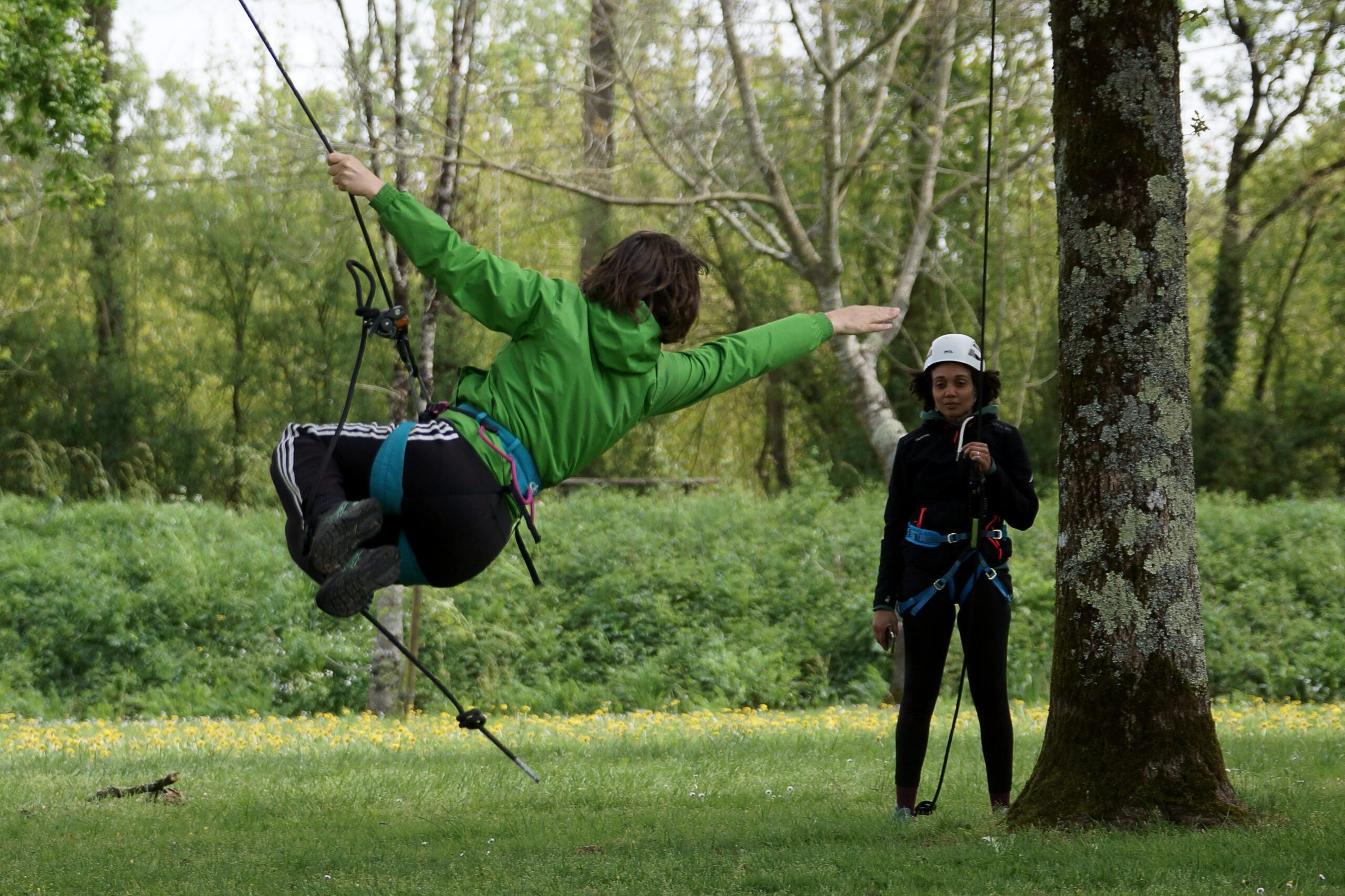 Lors d'un séminaire de team building, une personne portant une veste verte se balance sur une corde entre les arbres tandis qu'une autre, portant un casque blanc, se tient à proximité sur l'herbe.