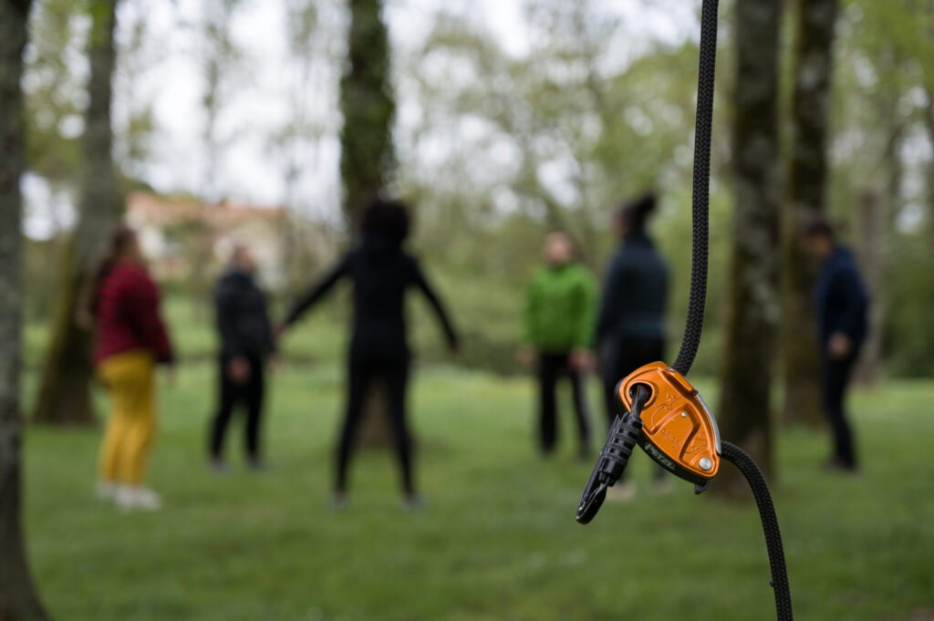 Un dispositif d'assurage orange attaché à une corde est mis au point, avec un groupe de personnes se tenant dans un arrière-plan flou parmi des arbres dans un environnement extérieur.