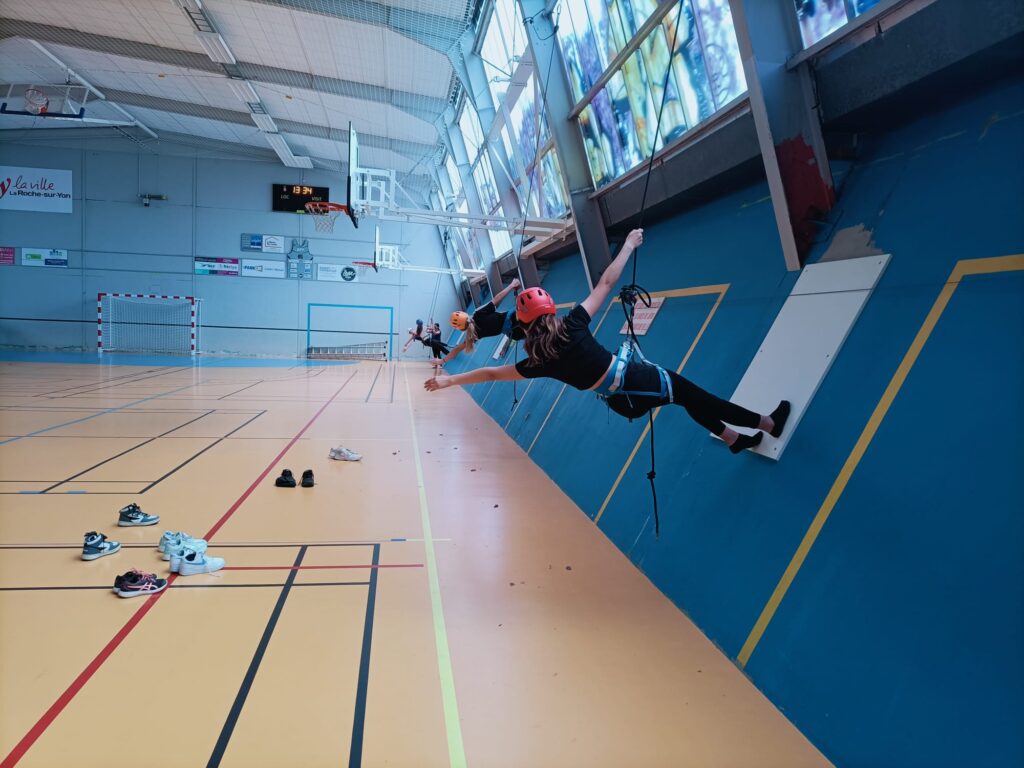 Deux personnes portant des casques et des harnais s'exercent à la danse-escalade sur un mur d'un gymnase intérieur ; des chaussures sont éparpillées sur le sol du gymnase à proximité.