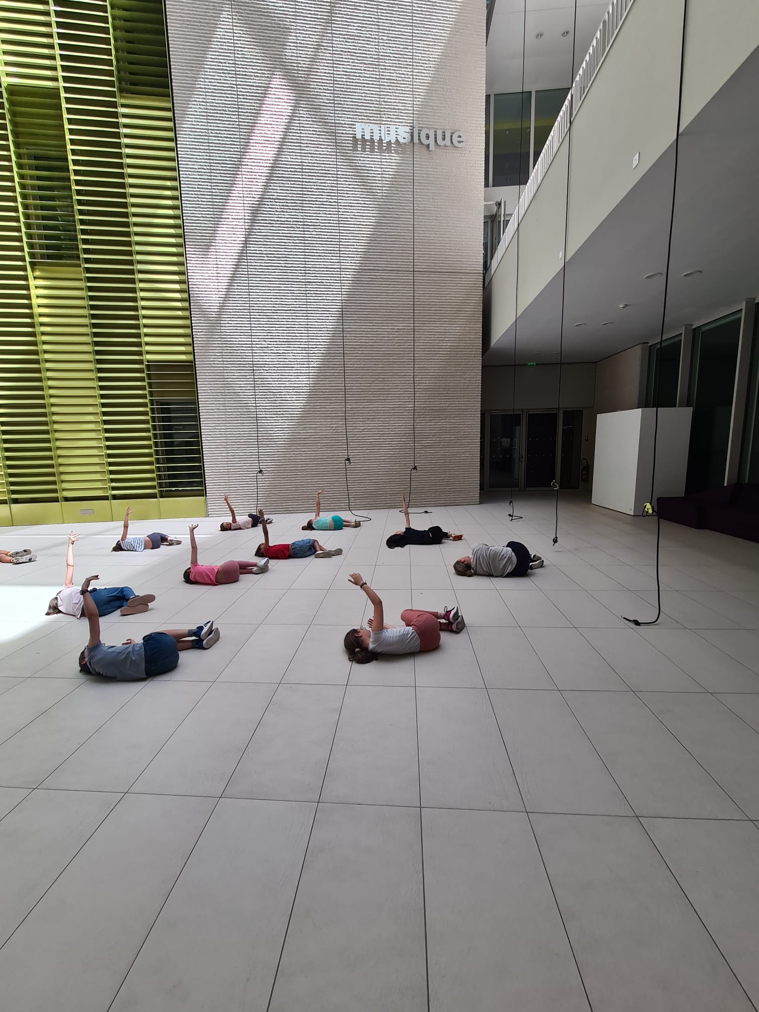 Un groupe de personnes, dont des enfants, est allongé sur le dos sur un sol carrelé à l'intérieur, chacun levant un bras vers des cordes suspendues dans une danse verticale. Le mot musique est visible sur le mur.