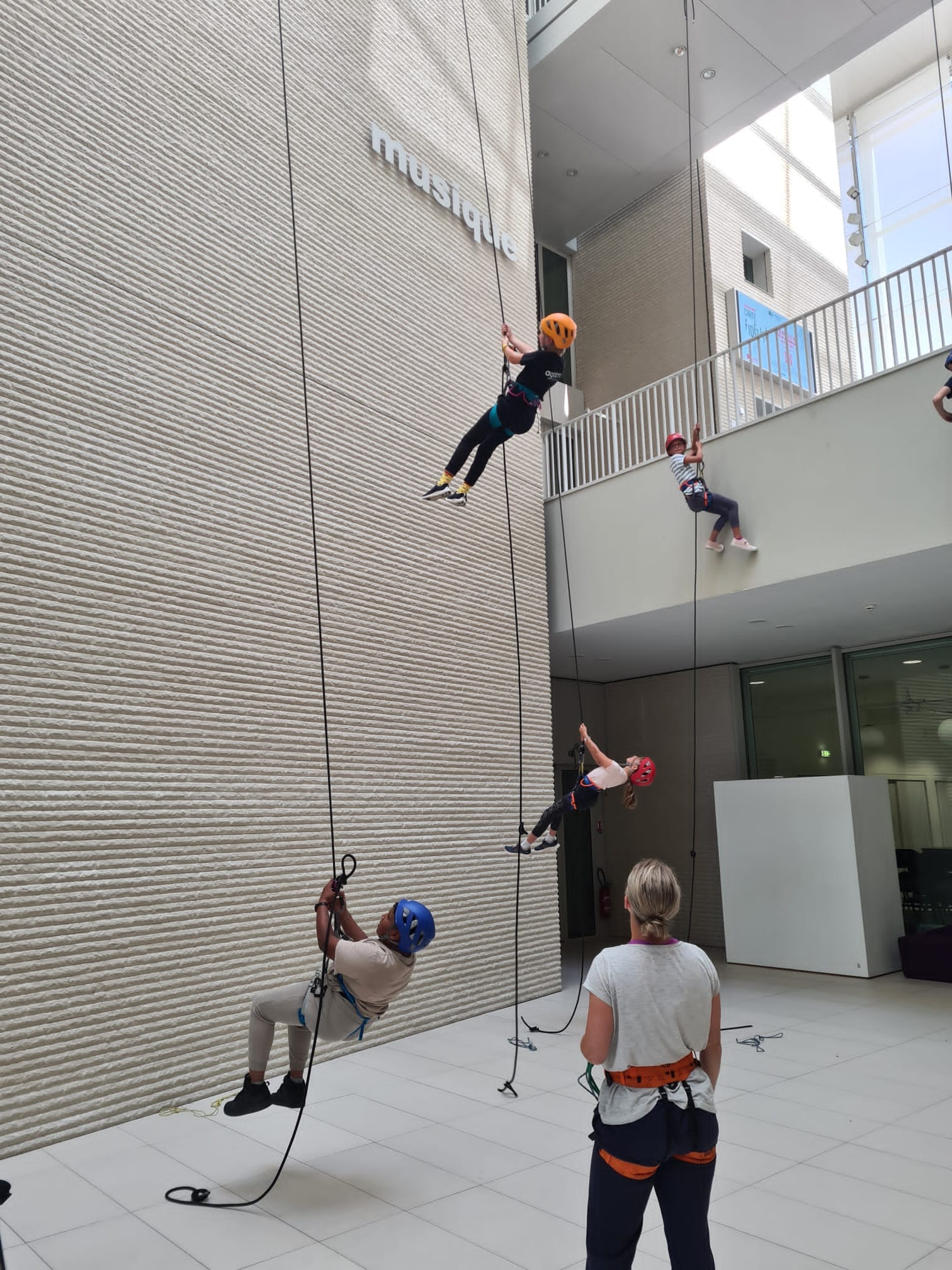 Quatre personnes, portant des casques et des harnais, grimpent et descendent en rappel à l'intérieur d'un bâtiment moderne pendant qu'un instructeur observe depuis le sol - une introduction passionnante à la danse verticale enfants.