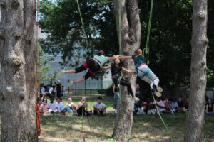 Trois personnes suspendues à des harnais se tendent l'une vers l'autre entre les arbres, exécutant une danse d'arbre, tandis qu'une troisième personne se tient entre elles ; un groupe de personnes du collège observe à l'arrière-plan.