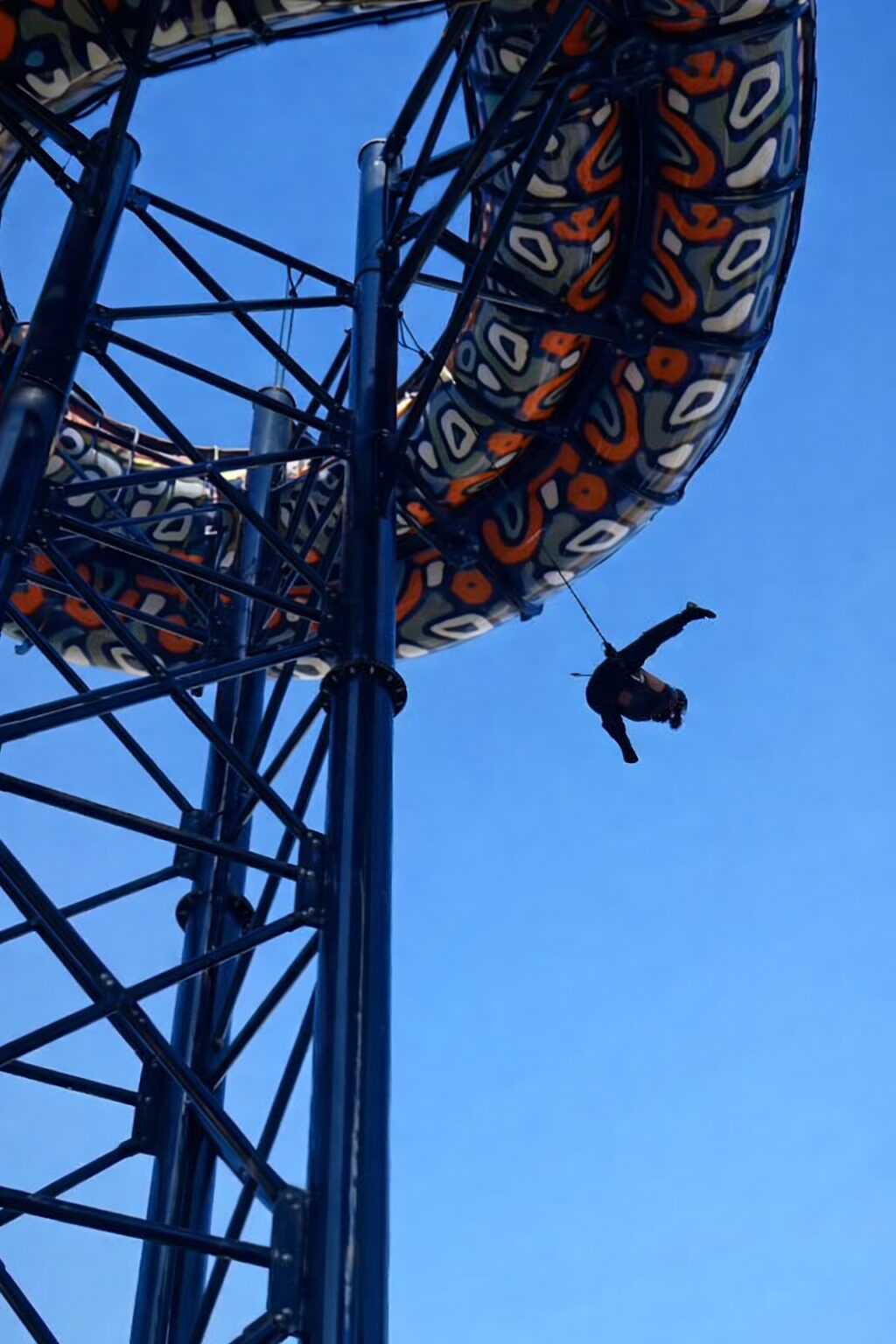 Une personne est suspendue à l'aide d'un harnais sous un grand tobogan sous un ciel bleu limpide.