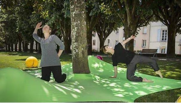 Deux personnes posent sur une surface verte déformée et ondulée qui déforme le sol autour d'un arbre dans un parc avec des bâtiments en arrière-plan.
