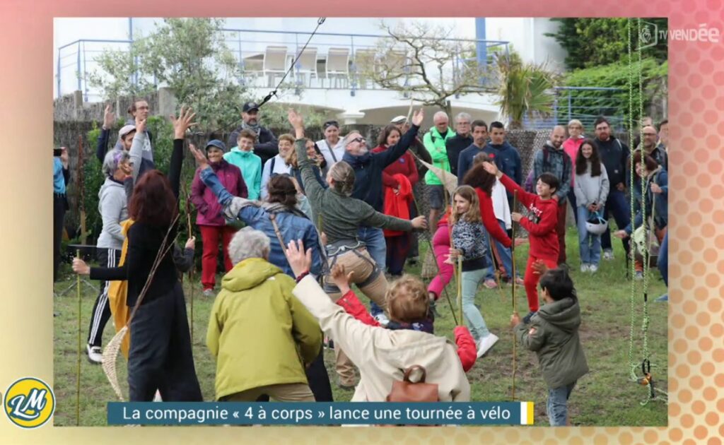 Un groupe de personnes, comprenant des enfants et des adultes, participe à une activité en plein air, les bras levés, sous le regard d'une foule sur un terrain herbeux.