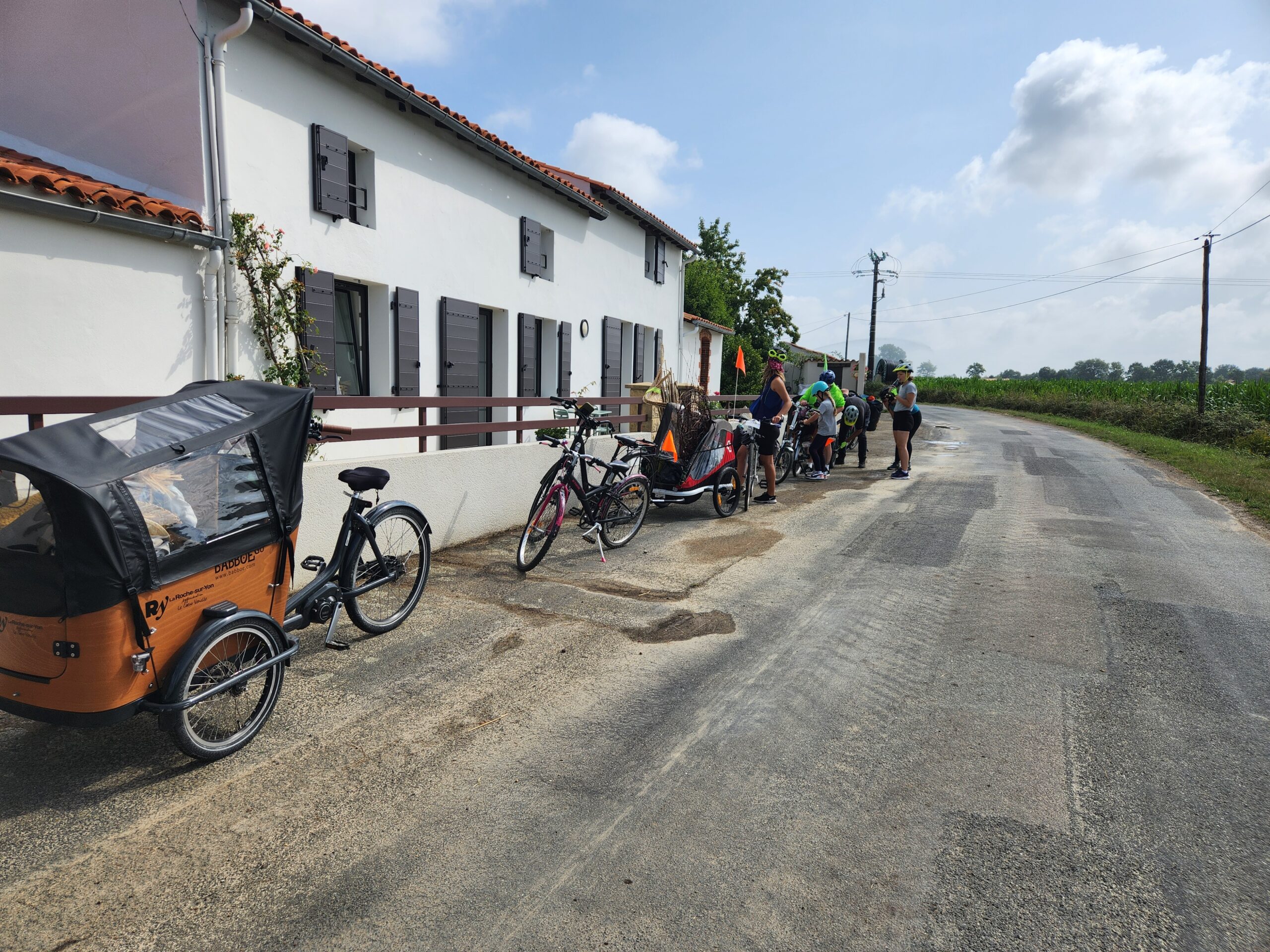 Un groupe de personnes se tient près d'une maison blanche au bord d'une route de campagne en Vendée, où plusieurs vélos, dont un vélo cargo, sont garés à proximité, ce qui laisse penser à un rassemblement en plein air.