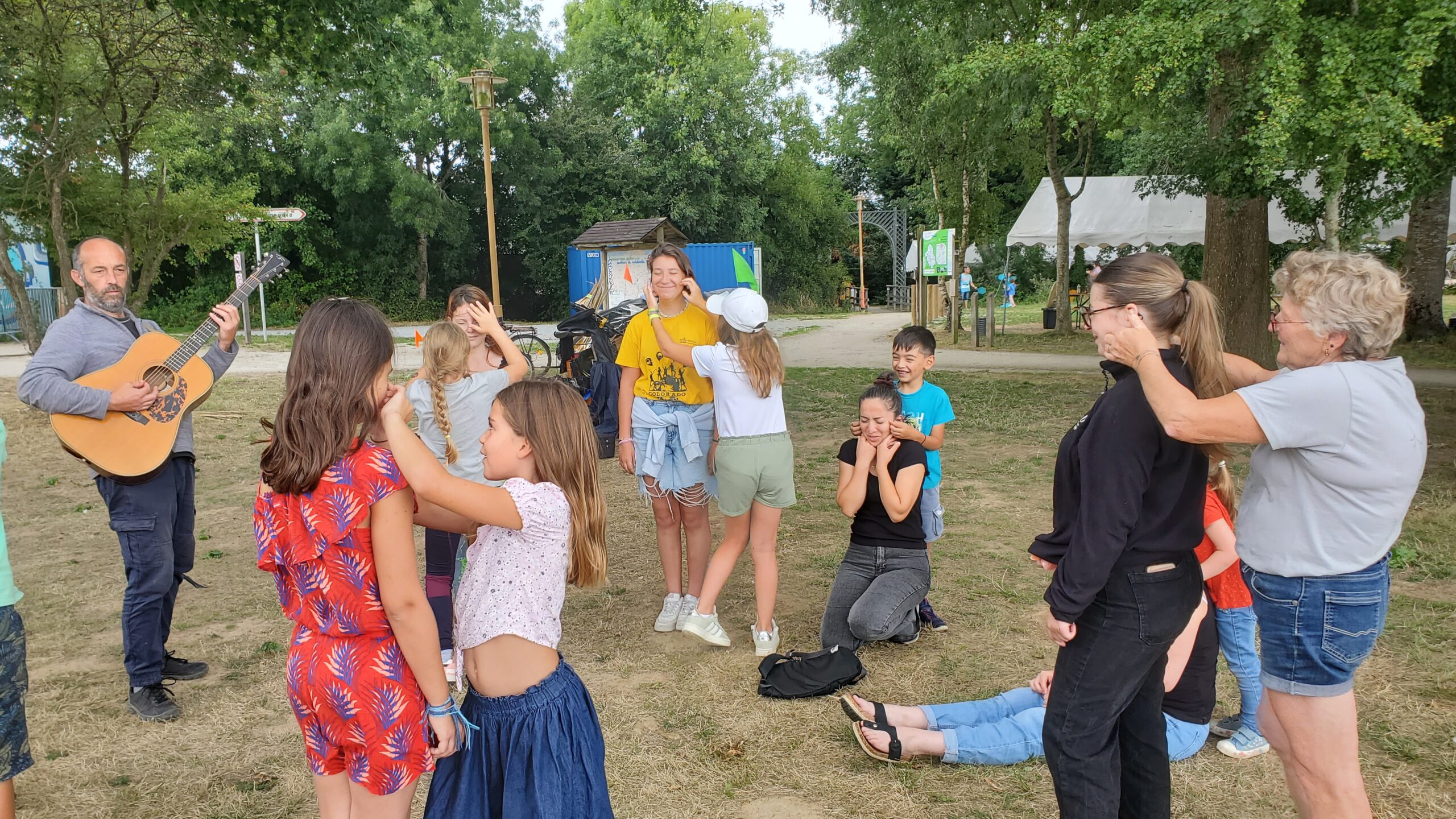 Un groupe d'enfants et d'adultes participent à un atelier de danse en extérieur, tandis qu'un homme joue de la guitare ; certaines personnes se couvrent les yeux, avec des arbres et des tentes visibles en arrière-plan.