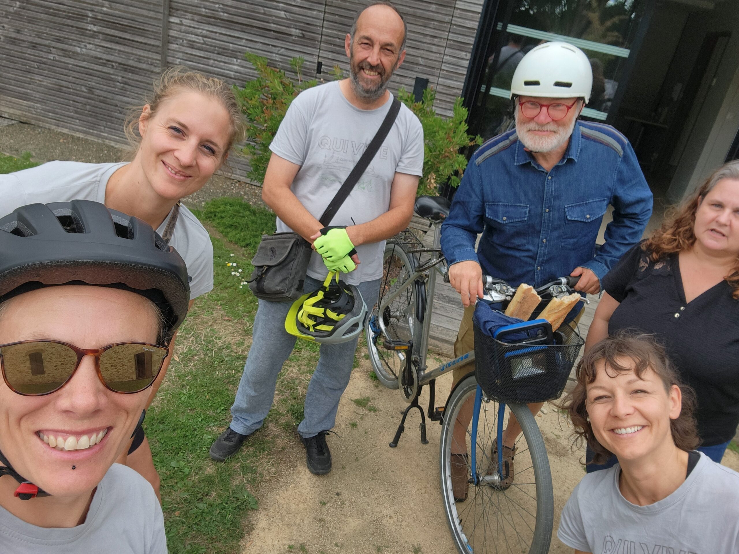 Cinq personnes se tiennent à l'extérieur, souriant pour la photo. Deux d'entre elles portent des casques de vélo, une autre tient un casque et des gants, et un homme se tient à côté d'un vélo