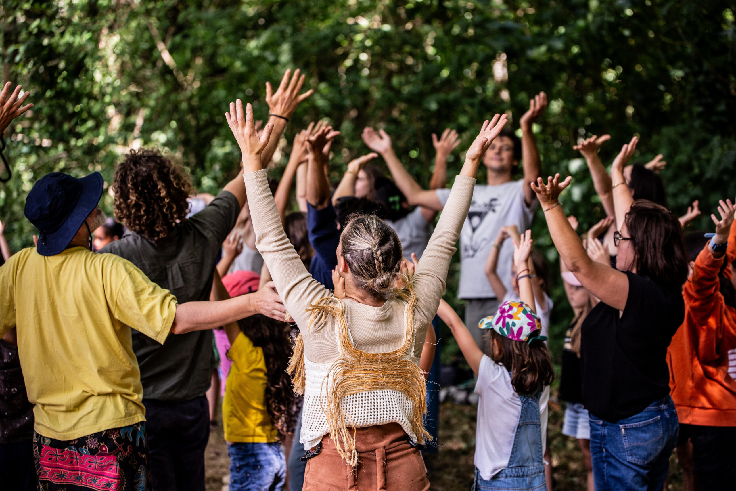 Un groupe de personnes, composé d'adultes et d'enfants, se tient debout en cercle à l'extérieur, les bras levés, profitant d'un spectacle en plein air au milieu des arbres et d'une végétation luxuriante.