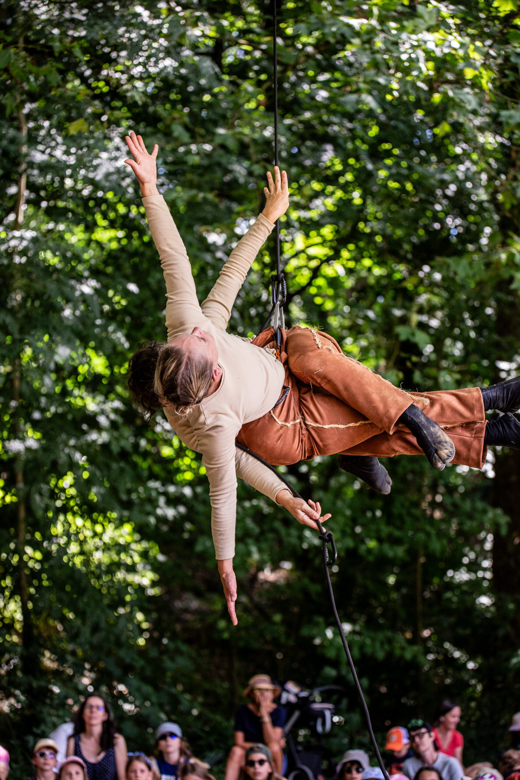 2 artistes vêtues d'un pantalon marron et d'une chemise beige est suspendu à un harnais, les bras tendus, suspendues à un arbre, sous le regard d'un public dans un spectacle de danse aérienne