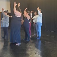Sept personnes se tiennent en rang sur la piste d'un studio de danse, le bras droit levé, face à un rideau noir, lors d'une séance d'atelier de danse contemporaine.