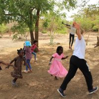 Un groupe d'enfants danse en plein air sur un terrain naturel bordé d'arbres, leurs rires résonnent dans la nature comme dans une danse joyeuse.