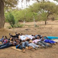 Un groupe d'enfants est allongé sur le dos sur une natte sous un grand arbre dans un espace extérieur sec et ouvert au Sénégal, entouré d'arbres et d'arbustes épars, profitant du moment après une séance animée de danse dehors.