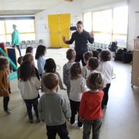 Un groupe de jeunes enfants se tient dans une salle de classe, participant à une activité de danse contemporaine école, face à un adulte qui semble donner des instructions ; deux autres adultes sont présents à l'arrière-plan.